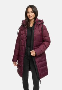 Mujer vestida con un abrigo de invierno largo y acolchado en color burdeos con capucha, una camiseta negra y jeans oscuros, de pie contra un fondo claro y sencillo.