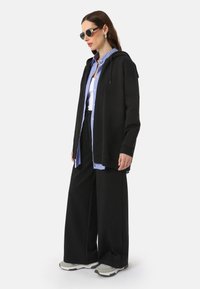 Black zip-up hoodie with a relaxed fit, layered over a blue and white striped shirt, paired with wide black pants and gray sneakers.
