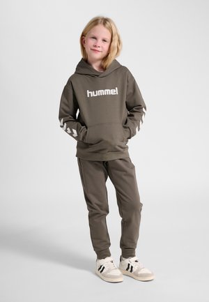 Barn med blondt hår iført en brun hættetrøje og matchende bukser med hvide sneakers, stående med hænderne i lommerne mod en ensfarvet baggrund.