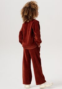 Brun fleece-träningsdräkt med avslappnad passform; har långa ärmar, rundad halsringning och vida byxor. Bärs med benvita sneakers.