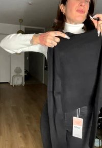 Personne tenant un pull noir avec une étiquette visible "ONLY", portant un col roulé blanc, debout dans une pièce avec un sol en bois.