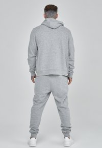 Felpa grigia con cappuccio e pantaloni della tuta abbinati in morbido tessuto. La felpa presenta un cappuccio e polsini a coste. L'outfit è completato da sneakers bianche.