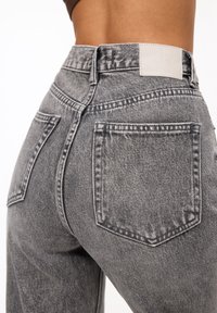 Jeans de denim gris de cintura alta con un lavado desvanecido, que presentan dos bolsillos traseros y una etiqueta en la cinturilla. Tejido texturizado, diseño casual.