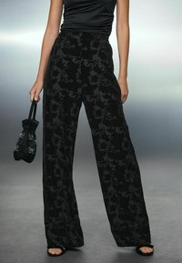 Pantaloni negri cu picioare largi, cu un model floral, dintr-un material texturat, asortați cu o poșetă mică din mărgele negre. Sandale vizibile pe picioare.
