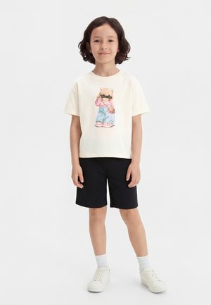 Enfant aux cheveux courts et foncés portant un t-shirt crème avec un motif d'ours, un short noir, des baskets blanches et des chaussettes blanches, debout devant un fond uni.