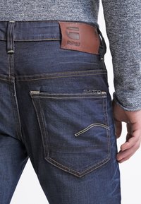Mörkblå denimjeans med en brun läderlapp märkt "RAW" vid midjan. Har gula sömmar och en ficka med dragkedjedetalj.