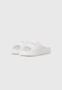 Sandales en caoutchouc blanches à semelle ouverte et talon découvert, dotées d'une semelle texturée et d'un logo "Lacoste" en violet embossé sur le côté extérieur.