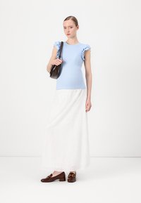Haut bleu clair avec des manches volantes, jupe maxi blanche avec un motif en œillets, mocassins marron et un sac à l'épaule sombre.