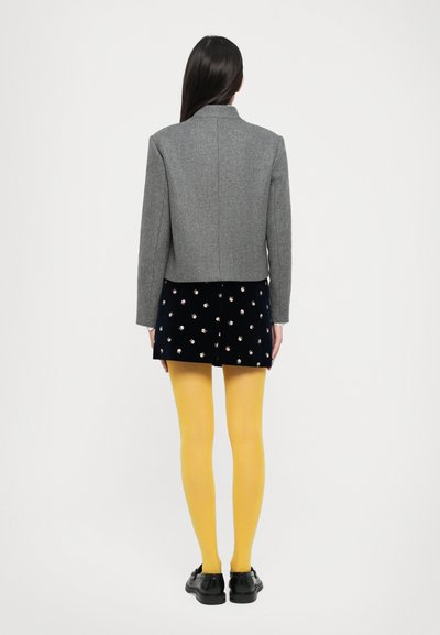 Veste cropped en mélange de laine grise avec un col haut, associée à une jupe courte navy à motifs floraux et des collants jaunes. Chaussures noires.