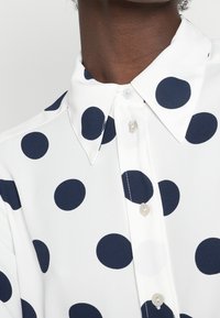 Zbliżenie na osobę noszącą białą koszulę w duże granatowe kropki, z kołnierzykiem w kształcie ostrego i białymi guzikami.