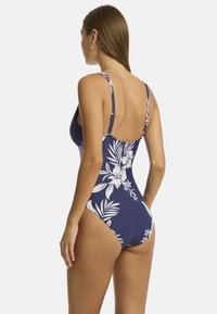 Mujer con un traje de baño floral azul marino, vista desde atrás, que muestra los patrones florales y tirantes ajustables, contra un fondo blanco liso.