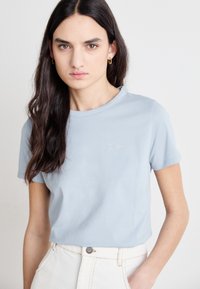 Light blue cotton t-shirt with short sleeves, round neckline, and a small silver embroidered logo on the chest, paired with white high-waisted pants.