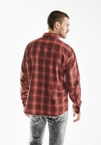 Červená károvaná košile s dlouhým rukávem a límcem na knoflíky. Košile má volnější střih a měkkou texturu látky. Viditelné detaily šití.