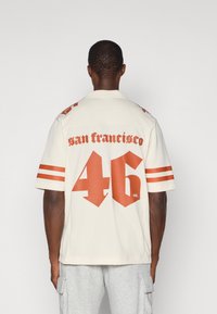 Krémové tri�čko s oranžovými pruhy a velkým textem "san francisco" a "46" na zadní straně. Krátké rukávy, volný střih, strukturovaná tkanina.