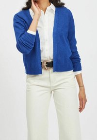 Modrý kardigan z pleteného materiálu, s výstřihem do V, otevřeným předním dílem a žebrovanými rukávy. Kombinován s béžovými širokými kalhotami a pásem.