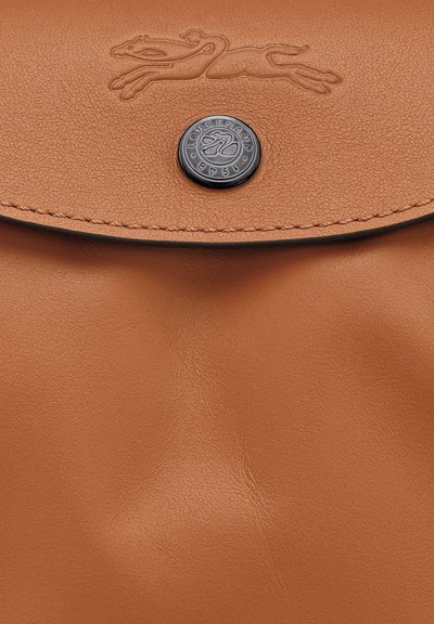 Tan leather bag flap with embossed horse logo and black circular button engraved with "Longchamp 1948" close-up.