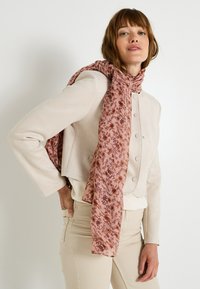 Veste courte en suède beige avec fermeture à bouton, accompagnée d'un foulard floral rose drapé autour du cou, portée avec un pantalon de couleur claire.