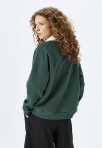 Przytulny, oversized zielony sweatshirt z miękkiego materiału, z prążkowanym wykończeniem na dole i rękawach. Kołnierz jest biały, co tworzy kontrast w projekcie.