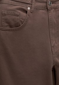 Gros plan sur le tissu marron du pantalon montrant la poche avant, la ceinture avec passant de ceinture, le bouton et les coutures détaillées.