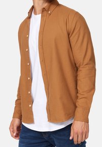 Braunes Hemd mit Knöpfen aus strukturiertem Stoff, ausgestattet mit einem Kragen, langen Ärmeln und einem abgerundeten Saum, kombiniert mit einem weißen T-Shirt darunter.