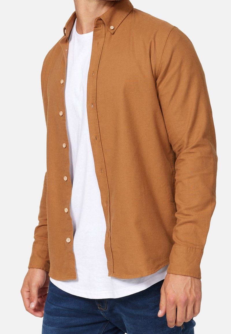 Braunes Hemd mit Knöpfen aus strukturiertem Stoff, ausgestattet mit einem Kragen, langen Ärmeln und einem abgerundeten Saum, kombiniert mit einem weißen T-Shirt darunter.