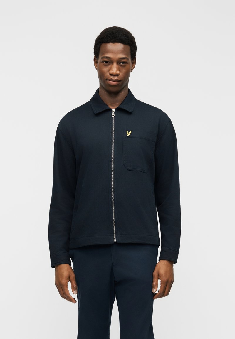 Νεαρός άνδρας φοράει μπλε σακάκι με φερμουάρ, με τσέπη στο στήθος και κίτρινο λογότυπο πουλιού, στέκεται μπροστά σε απλό ανοιχτό γκρι φόντο.