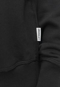 Svart sweatshirttyg som visar en närbild av sömmen med en liten vit etikett märkt "ORIGINALS™." Slät textur och figurnära design.