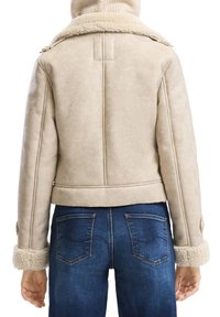 Persoon in een beige schapenvachtjas met opgerolde cuffs en kraag, gecombineerd met donkerblauwe spijkerbroek, gezien vanuit de achterkant.