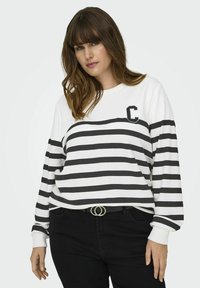 Sudadera blanca con rayas horizontales negras y un pequeño logotipo "C" en el lado izquierdo del pecho. El modelo la lleva con pantalones negros y un cinturón plateado.