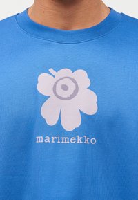 Sinine särk, millel suur valge lille graafika ja all kirjas "marimekko", mis asub lille all rinnal keskel.