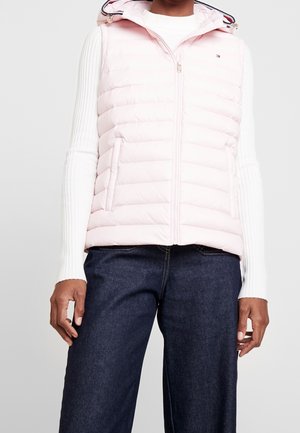 Person iført en lyserød quiltet ærmeløs hættetrøje over en hvid ribbet langærmet top og mørkeblå jeans.