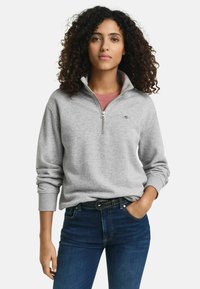 Grijze rits sweatshirt met een hoge kraag, gemaakt van zacht materiaal, met ribgebreide manchetten en een klein logo. Gepaard met donkere jeans en een riem.