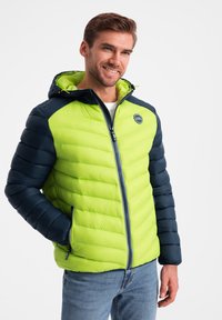 Vaatteet pehmustetut takki kirkkaan limevihreällä vartalolla ja tummansinisin hihoilla, jossa on huppu, vetoketju ja logopatch rinnan alueella.