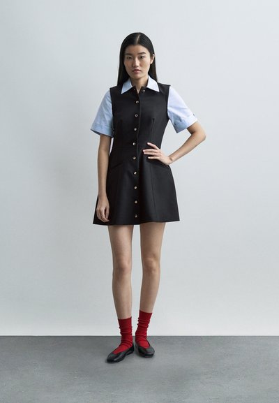 Γυναίκα στέκεται με το χέρι στη μέση, φορώντας μαύρο φόρεμα με κουμπιά πάνω από κοντομάνικο πουκάμισο, κόκκινες κάλτσες και μαύρα παπούτσια μπαλαρίνας, μπροστά σε απλό φόντο.