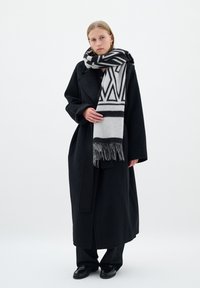 Lungo cappotto nero con silhouette oversize, abbinato a una sciarpa grigia a fantasia con disegni geometrici neri e bordi frangiati. Pantaloni e scarpe neri.