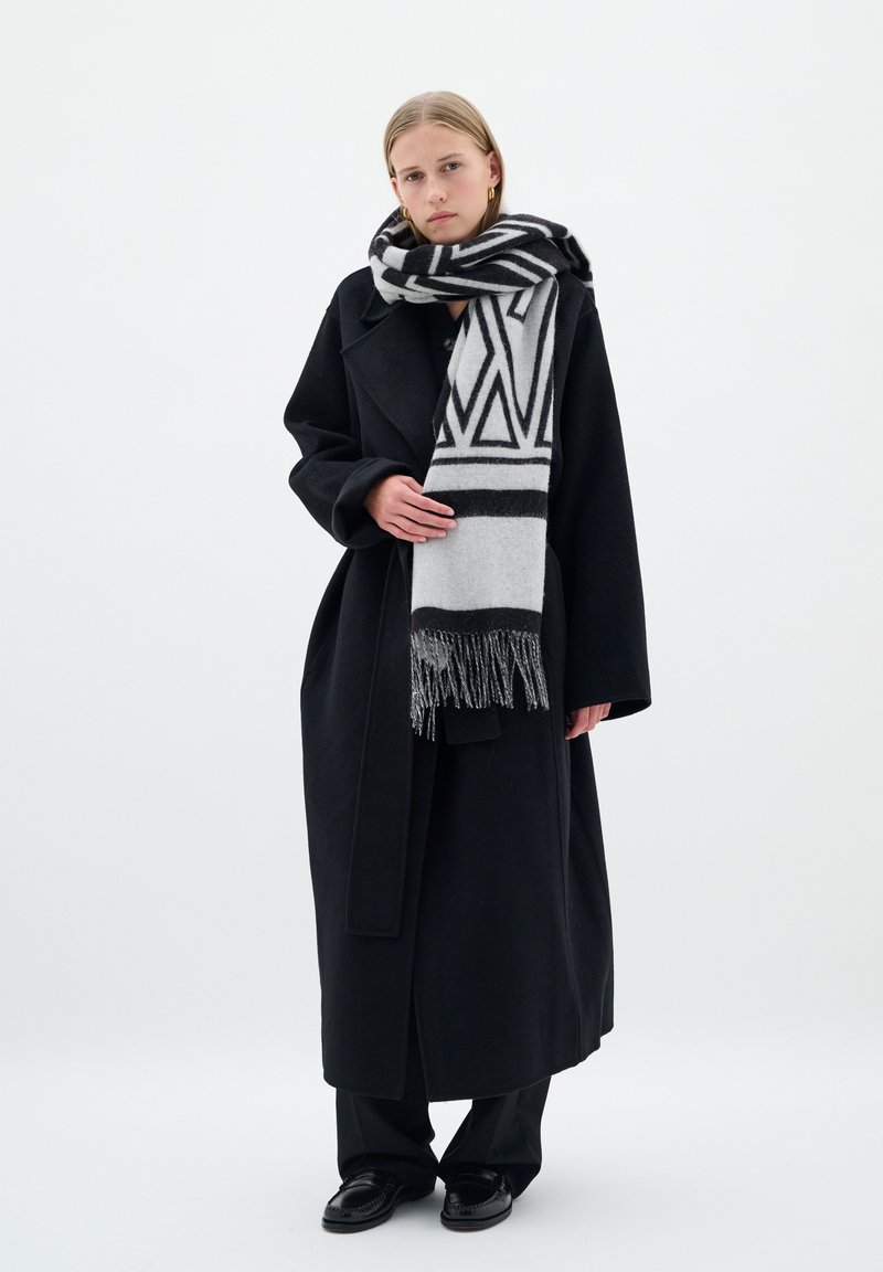 Lungo cappotto nero con silhouette oversize, abbinato a una sciarpa grigia a fantasia con disegni geometrici neri e bordi frangiati. Pantaloni e scarpe neri.