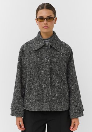 Femme portant une veste grise à motif chevrons avec un grand col et un bouton, des lunettes de soleil rectangulaires teintées et un pantalon noir.