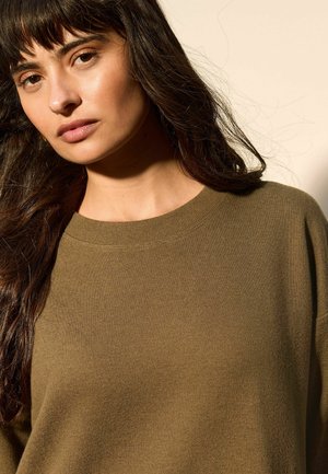 Mujer con cabello largo y oscuro, vistiendo un suéter de punto verde oliva, mirando ligeramente hacia un lado contra un fondo beige.