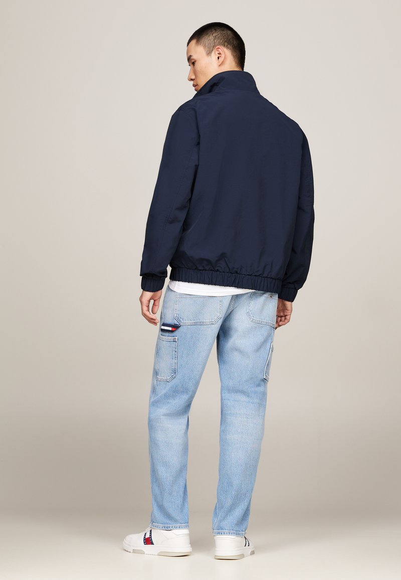 Veste bomber bleu marine avec col montant, poignets élastiques et ourlet, associée à un jean bleu clair et des baskets blanches avec des accents rouges.