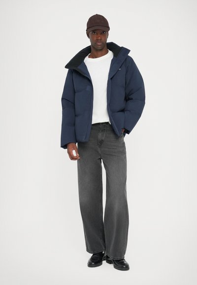 Navy puffer jacket with black collar, white T-shirt underneath, paired with gray wide-leg jeans and black shoes. Man wearing a brown cap.