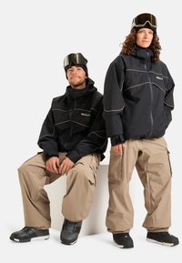 Du žmonės vilki juodus švarkus su smėlio spalvos akcentais, derinamus su laisvais smėlio spalvos kelnių ir sniego batais. Švarkai turi aukštą apykaklę ir užtrauktukus.