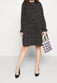 Svart långärmad klänning med vita prickar, strukturerad tyg, rynkade axlar och hög halsringning; matchad med en mönstrad handväska.