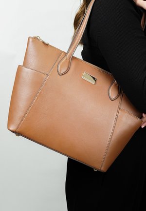 Sac fourre-tout en cuir tan avec surface texturée, double poignées, fermeture éclair et plaque au nom dorée. Dispose d'une poche latérale pour un rangement supplémentaire.