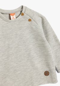 Jasnoszary sweter z prążkowanego materiału, z dwoma brązowymi zatrzaskami na ramieniu, małą okrągłą metką logotypu w lewym dolnym rogu oraz długimi rękawami. Miękka tekstura.