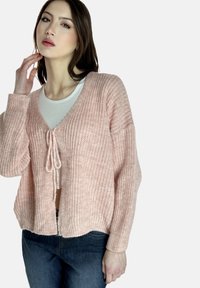 Sanft rosa gestrickte Strickjacke mit V-Ausschnitt und Bindeschluss. Verfügt über übergroße Ärmel und einen leicht verkürzten Saum, kombiniert mit einem weißen Oberteil und Jeans.