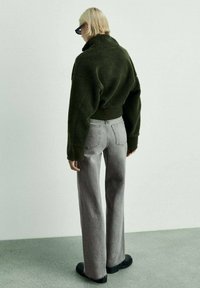 Zeleno prevelik flis jakna z visokim ovratnikom, v kombinaciji s svetlo sivo širokimi hlačami in črnimi ravnimi čevlji. Gladek, teksturiran material.