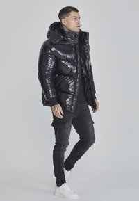 Veste doudoune glossy noire avec une grande capuche, fermeture éclair à l'avant et poignets côtelés, associée à un pantalon cargo noir et des baskets blanches.