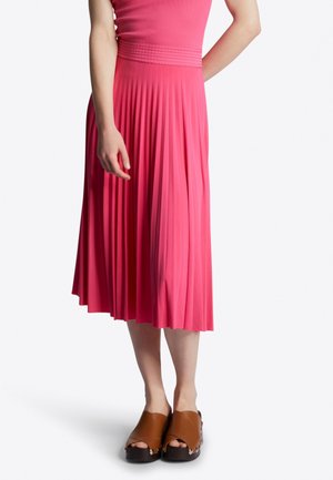 Mujer con falda midi plisada rosa, top a juego rosa y sandalias de plataforma marrones con tachuelas metálicas sobre fondo blanco.