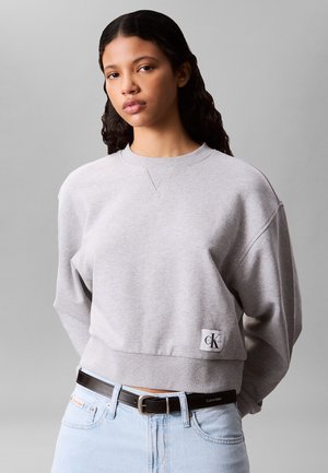 Fiatal nő hosszú sötét hajjal világosszürke Calvin Klein pulóvert és világoskék farmert visel fekete Calvin Klein övvel.