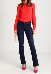 Röd långärmad blus, mörka utsvängda jeans, rosa högklackade skor. Modellen håller en väska i konstpäls och visar upp ett slätt tyg och en skräddarsydd passform.